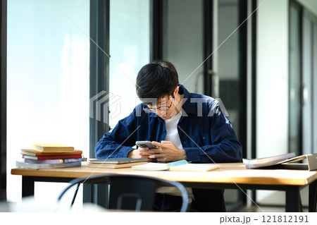 Student man using a smartphone at wooden desk surrounded by study materials 121812191