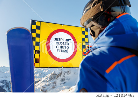 Advanced skier wear blue gear looking at multilingual warning sign "Closed" on snowy mountain resort under clear blue skies. Safety in winter mountain sports concept. Avalanche weather risk danger 121814465