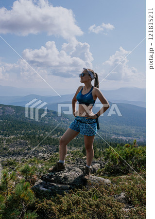 Woman in blue shorts and a sports bra stands on a stone against the backdrop of a summer landscape in the mountains Woman in blue shorts and a sports bra stands on a stone against the backdrop of a summer landscape in the mountains 121815211