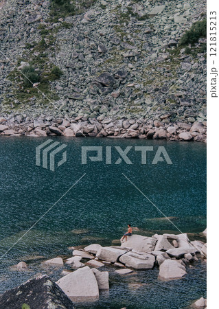 Man sits on large stone protruding from the water of emerald colored mountain lake. Natural landscape, summer hiking day 121815213