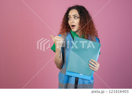Front view of shocked, surprised woman with open mouth looking aside, pointing, holding folders. Isolated on pink studio background. 121816403