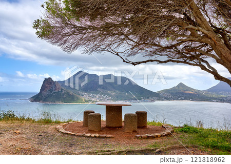Public bench overlooking the ocean and mountain looks beautiful out in nature. Landscape of four empty chairs and a table with a tree giving them shade offers a relaxing and romantic outdoor sitting Public bench overlooking the ocean and mountain looks beautiful out in nature. Landscape of four empty chairs and a table with a tree giving them shade offers a relaxing and romantic outdoor sitting 121818962