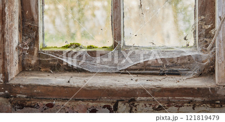 Abandoned, dirty and dusty window covered in spiderwebs in empty house from poverty and economic crisis. Old, damaged and weathered wooden windowsill and wood frame rotting from dampness and neglect 121819479