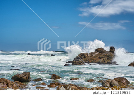 Big waves splashing and breaking on the coastline. Turbulent sea with rough tides from strong winds crashing onto beach boulders with a blue sky background. Rocky coast in Camps Bay, South Africa 121819652