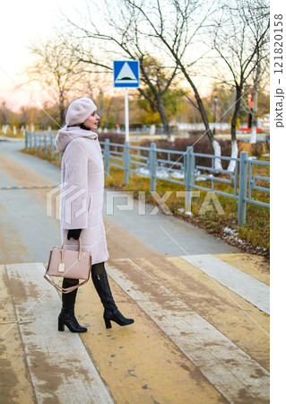 A Middle-Aged Woman in a Light Autumn Coat and Hat Stands Thoughtfully on the Sidewalk, Embracing the Crisp Fall Air and Reflecting on Life's Journey Amidst the Golden Leaves and Soft Light of the 121820158