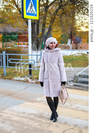 A Middle-Aged Woman in a Light Autumn Coat and Hat Stands Thoughtfully on the Sidewalk, Embracing the Crisp Fall Air and Reflecting on Life's Journey Amidst the Golden Leaves and Soft Light of the 121820159