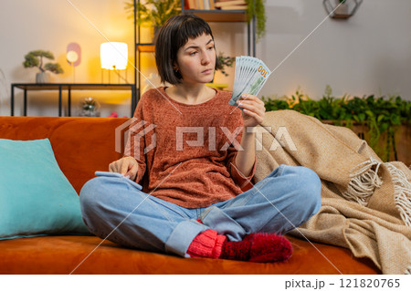 Woman counting money dollar banknotes savings earnings using smartphone calculator on sofa at home 121820765