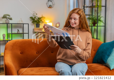 Woman accountant working with financial graph documents reading reports of income on sofa at home 121821412