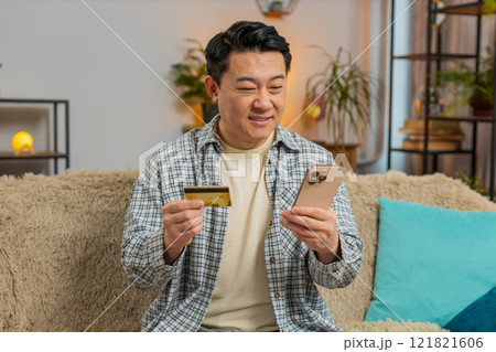 Happy middle-aged man using credit bank card and smartphone while shopping online sitting on sofa Happy middle-aged man using credit bank card and smartphone while shopping online sitting on sofa 121821606