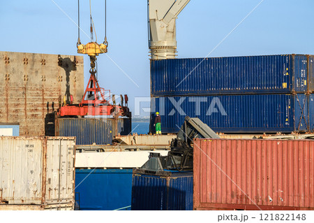 Sea port worker in safety gear supervises loading or unloading of cargo containers at busy shipping terminal. Cranes and container stacked, sunny day, clear blue sky, deck and ship-to-shore crane Sea port worker in safety gear supervises loading or unloading of cargo containers at busy shipping terminal. Cranes and container stacked, sunny day, clear blue sky, deck and ship-to-shore crane 121822148