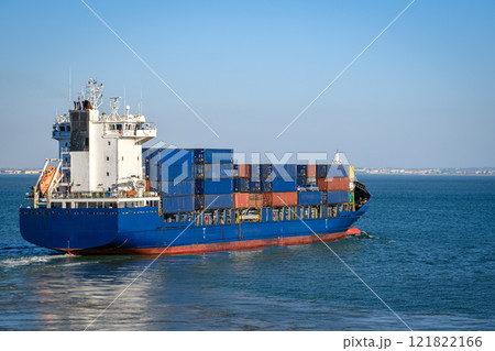 Modern blue bulk carrier loaded with stacked shipping containers navigating harbor near sea port. Global trade, maritime logistics, cargo, international freight. Modern blue bulk carrier loaded with stacked shipping containers navigating harbor near sea port. Global trade, maritime logistics, cargo, international freight. 121822166