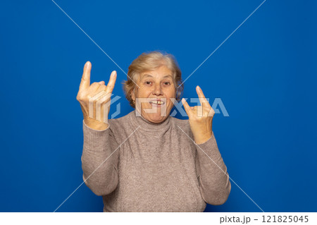 Caucasian woman about 70 years old wearing a dark beige sweater making a rock on sign symbol with hands up smiling broadly, isolated over blue background. Caucasian woman about 70 years old wearing a dark beige sweater making a rock on sign symbol with hands up smiling broadly, isolated over blue background. 121825045
