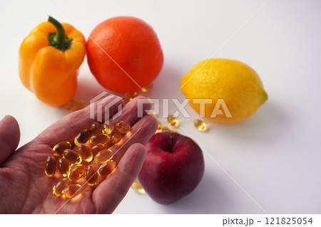 A hand holding several shiny golden capsules is extended in front of a vibrant arrangement of a yellow bell pepper, an orange, a lemon, and a red apple on a white background. A hand holding several shiny golden capsules is extended in front of a vibrant arrangement of a yellow bell pepper, an orange, a lemon, and a red apple on a white background. 121825054