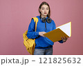 Front view of pretty schoolgirl standing with open mouth, looking at camera. Brunette female holding yellow open folder and rucksack, wearing casual clothes. Concept of youth. 121825632