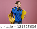 Front view of pretty brunette girl standing, touching ear, listening. Beautiful student with earphones, holding yellow rucksack and folder. Isolated on pink studio background. 121825636