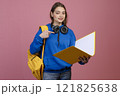 Front view of pretty schoolgirl standing, holding yellow backpack and folder. Young female student reading, looking at camera, smiling. Isolated on pink studio background. 121825638
