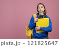 Front view of pretty female with rucksack and earphones standing with closed eyes. Beautiful schoolgirl wearing blue khudi, holding folder. Isolated on pink studio background. 121825647