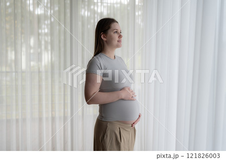 Portrait of pregnant gen z woman standing near window holds hands on her belly at home. Pregnancy, motherhood, expectation and tenderness concept 121826003