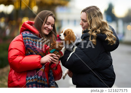 Two friends enjoying winter stroll while playing with a puppy in a festive outdoor setting Two friends enjoying winter stroll while playing with a puppy in a festive outdoor setting 121827575