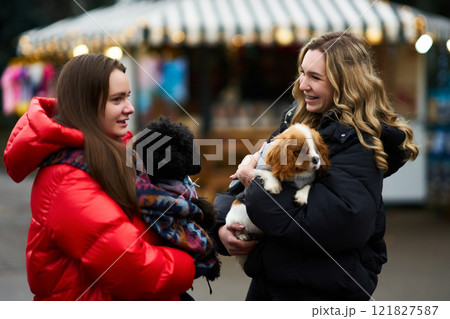 Two friends enjoy a winter day outside while holding small dogs at a festive outdoor market Two friends enjoy a winter day outside while holding small dogs at a festive outdoor market 121827587