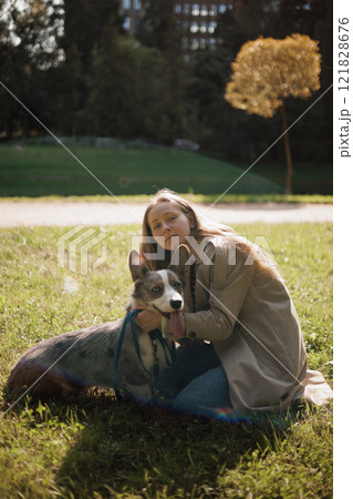owner walks on lawn in park with corgi dog. beautiful girl with long hair in beige raincoat. animal owner walks on lawn in park with corgi dog. beautiful girl with long hair in beige raincoat. animal 121828676