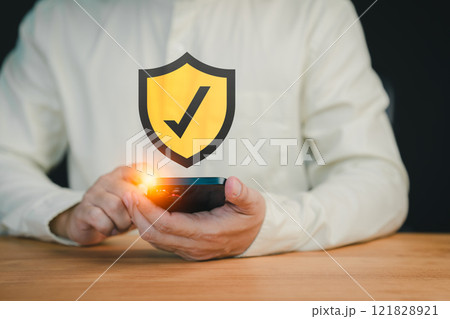 Close-up of a person holding a smartphone with a glowing shield icon featuring a check mark, symbolizing mobile security, data protection, and secure online verification. Close-up of a person holding a smartphone with a glowing shield icon featuring a check mark, symbolizing mobile security, data protection, and secure online verification. 121828921