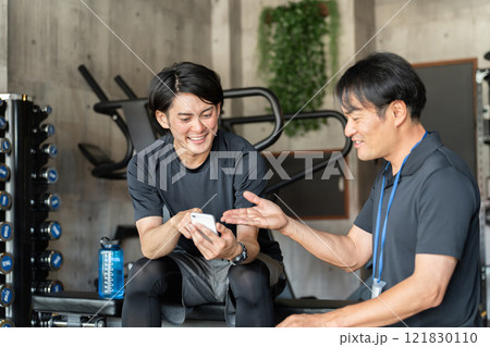 ジム・スポーツジムで会員男性にスマホでお得情報を案内する男性トレーナー 121830110