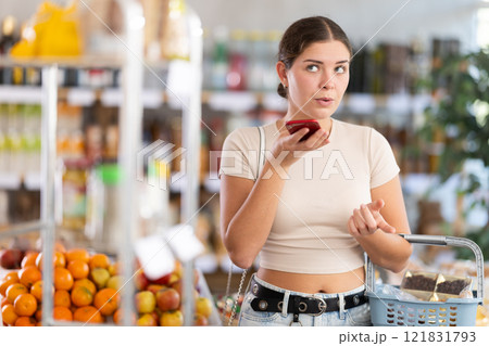 European girl with basket of groceries in hands stands in store and dictates audio message European girl with basket of groceries in hands stands in store and dictates audio message 121831793