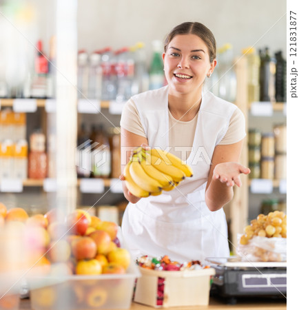 Young woman seller in store is standing near fruit counter, selling bananas. 121831877