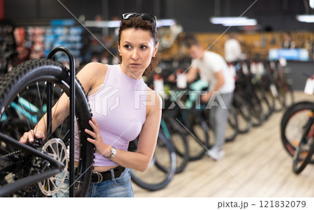 Young woman selects bicycle in sporting goods store, checking wheel quality Young woman selects bicycle in sporting goods store, checking wheel quality 121832079