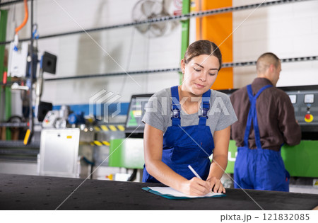Smiling female factory worker with clipboard standing in production workshop 121832085