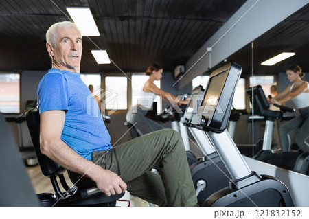 Elderly man, along with other people, is engaged on an bike in gym 121832152