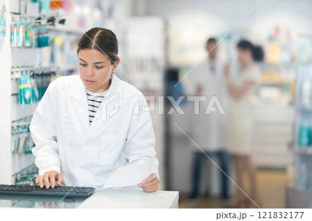 Attentive female pharmacist looking at computer in chemist shop with large assortment 121832177
