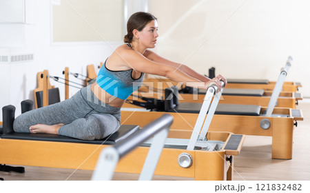 Woman doing pilates exercises lying on pilates workout machines in gym 121832482