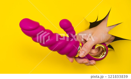 Female hand with magenta vibrator sticking out from yellow paper background. Copy space. Female hand with magenta vibrator sticking out from yellow paper background. Copy space. 121833839