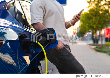 Man charges his electric vehicle while using a mobile phone in the street at sunset. Man charges his electric vehicle while using a mobile phone in the street at sunset. 121834292