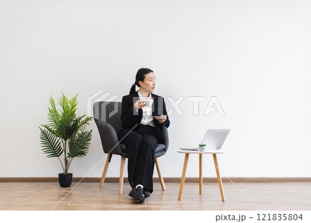 Asian businesswoman seated with coffee in minimalist office setting. Woman in professional suit working on laptop, creating calm business environment. Modern, clean design emphasizes productivity. 121835804