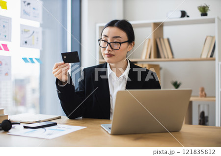 Asian business woman using credit card for online payment at office desk. Young female working on laptop, efficient and focused. 121835812
