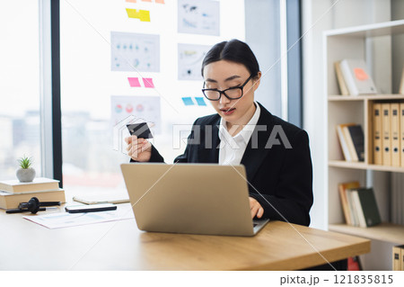 Asian business woman using credit card for online payment at office desk. Young female working on laptop, efficient and focused. 121835815
