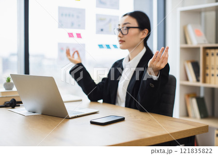 Asian businesswoman in her 30s meditating in lotus position at desk during break, promoting work-life balance. 121835822