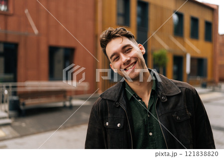 Cheerful young man wearing leather jacket and dark green shirt smiling looking at camera while standing in front of modern buildings in urban environment. Concept of modern city lifestyle. 121838820