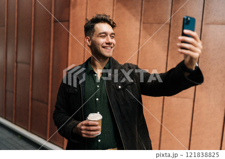 Cheerful man making a video call with smartphone while holding coffee cup, standing on background of modern building facade in urban environment. Concept of modern communication and urban lifestyle. 121838825