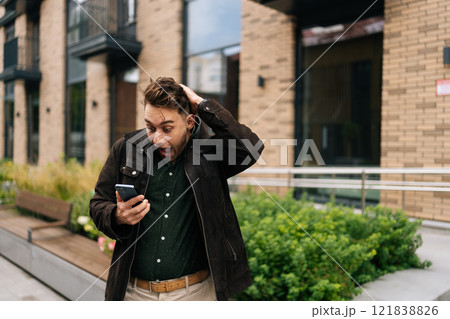 Surprised young man standing in urban street, holding smartphone and reacting with astonishment to some unexpected news just received, looking to phone screen with happy excited expression. 121838826