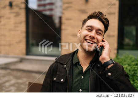 Laughing young man sitting on bench in city street, smiling and engaging in cheerful phone conversation while enjoying vibrant urban atmosphere and natural light around. Concept of city lifestyle. Laughing young man sitting on bench in city street, smiling and engaging in cheerful phone conversation while enjoying vibrant urban atmosphere and natural light around. Concept of city lifestyle. 121838827