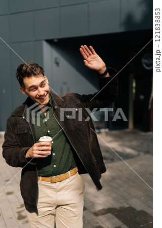 Vertical portrait of friendly young man dressed in casual attire, holding coffee cup, waving cheerfully at someone while walking along city street on sunny spring day. Concept of urban lifestyle. 121838853