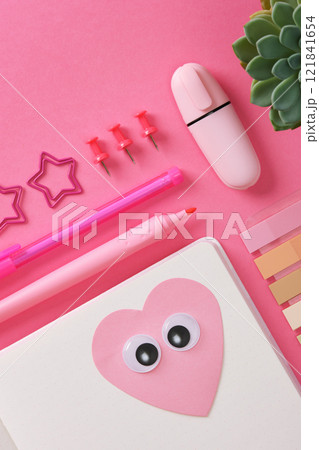 Heart-shaped sticker with eyes glued on the open page of a notepad. Pink themed background with stationery, paper clips, notepad, pencil, pen. Space for your text. Heart-shaped sticker with eyes glued on the open page of a notepad. Pink themed background with stationery, paper clips, notepad, pencil, pen. Space for your text. 121841654