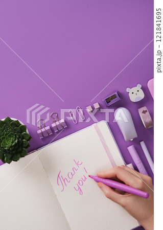A hand writes the words Thank you in English in a notebook. Against a background of purple stationery and heart shaped notes. A place for your text. A hand writes the words Thank you in English in a notebook. Against a background of purple stationery and heart shaped notes. A place for your text. 121841695