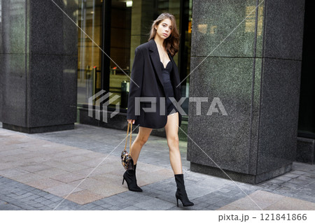 Beautiful Young Brunette Woman Walking City Street Near Modern Building, Stylish Fashion Model Wearing elegant outfit Oversized Blazer, Mini Dress, Small Handbag and Black Heeled Boots. Street style Beautiful Young Brunette Woman Walking City Street Near Modern Building, Stylish Fashion Model Wearing elegant outfit Oversized Blazer, Mini Dress, Small Handbag and Black Heeled Boots. Street style 121841866