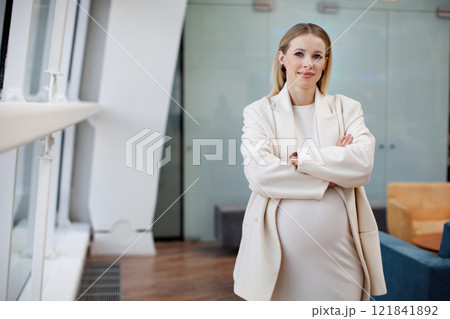 Beautiful, confident pregnant blonde businesswoman in stylish and fashionable clothing a white dress and blazer looking at the camera in a corporate office setting 121841892