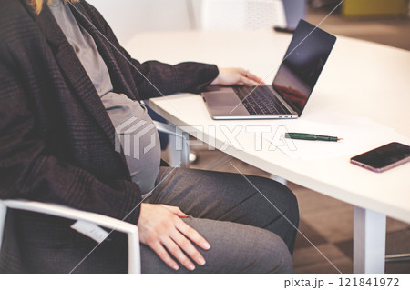 Close up Pregnant woman working on laptop at white desktop. Cropped pregnant businesswoman typing on laptop while sitting at her working place in office 121841972